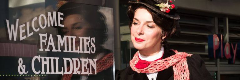 slide: Mary Poppins (Sara-Jeanne Hosie) smiling beside sign that reads “Welcome Families and Children”