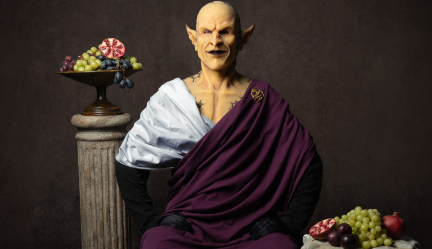 The goblin MUG, dressed in a burgundy and white robe, with bouquets of flowers in the back and foreground.